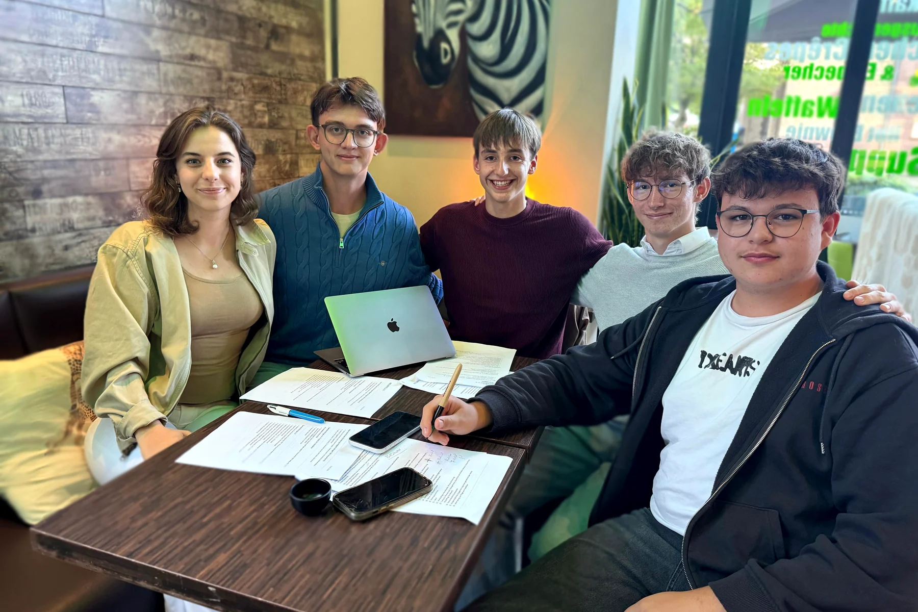 Fünf junge Menschen sitzen an einem Cafétisch, vor sich ausgedruckte Satzungsentwürfe, Stifte und einen Laptop.